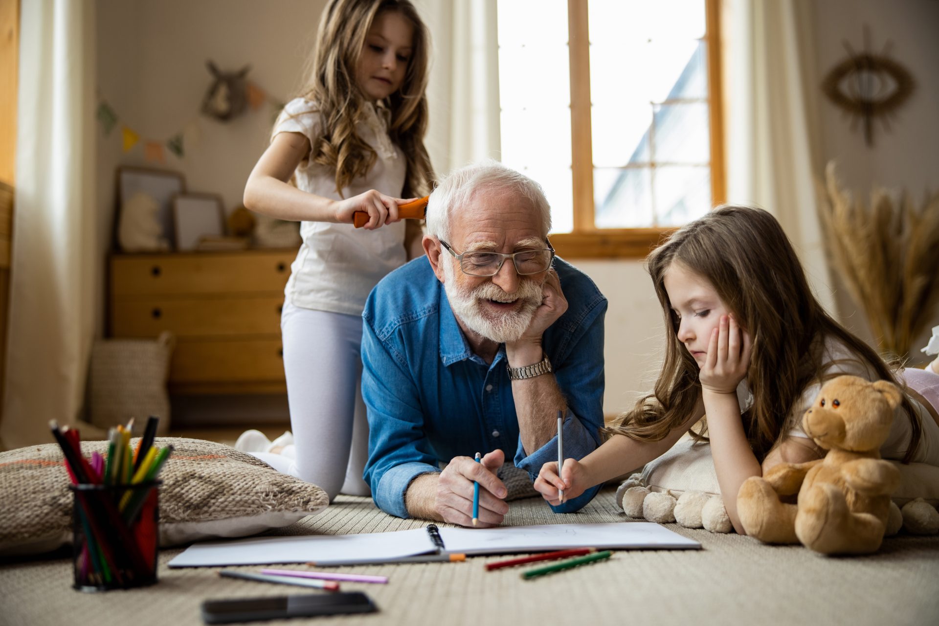Positive grandpa lying on the floor with colorful pencils and a careful granddaughter combing his hair