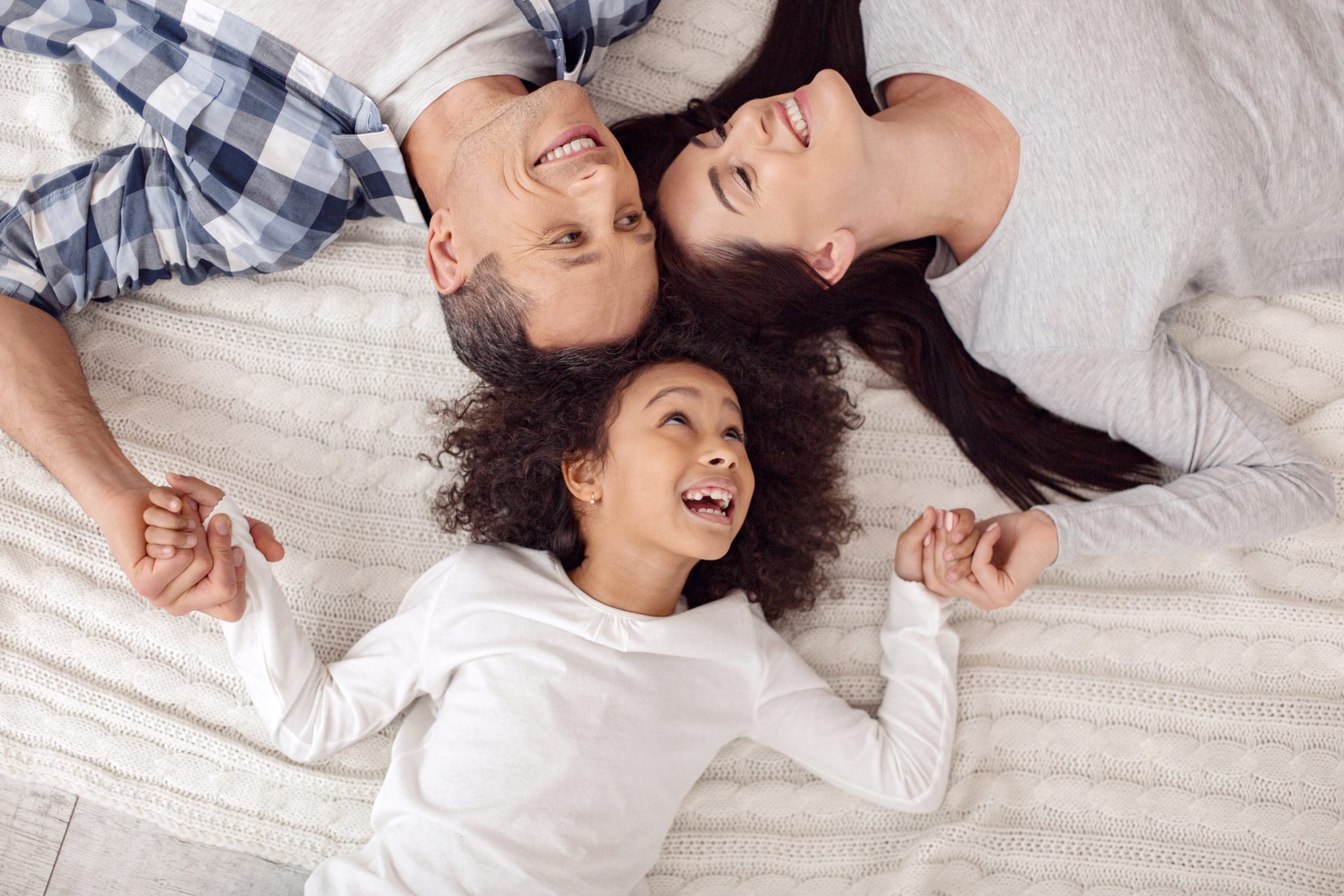 Relaxing. Adorable joyful curly-haired girl smiling and lying with her parents on bed and they looking at each other