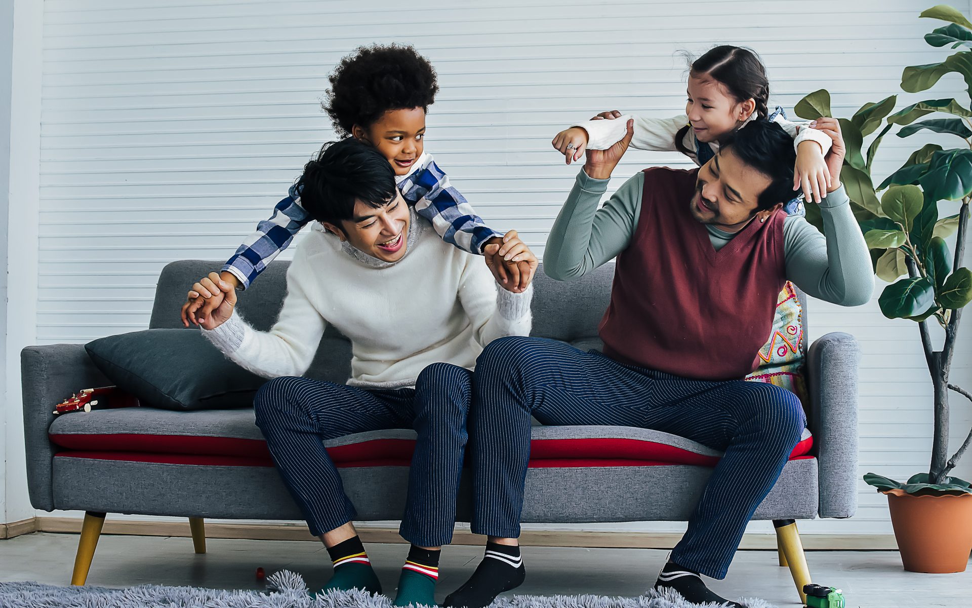 Male gay taking care adopted children who are happy diverse little Caucasian girl and African boy, hugging, playing toys together with fun, sitting on sofa in living room at home. LGBT, kids Concept