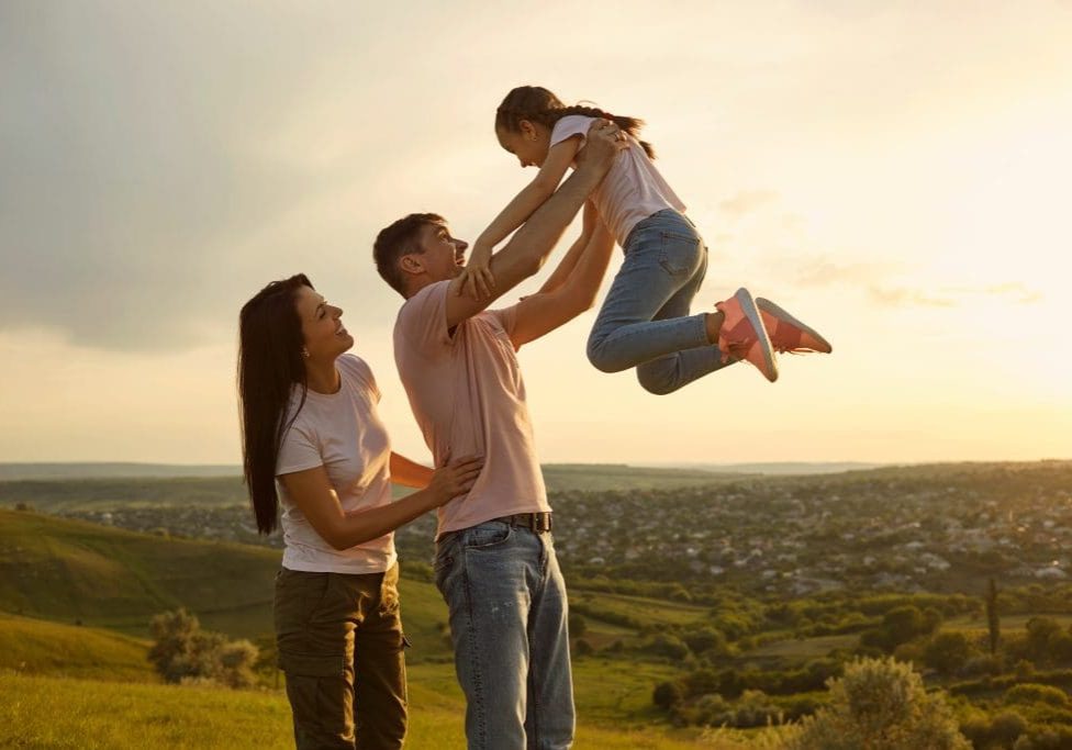 Joyful young family having fun with their daughter in mountains at sunset, blank space. Father lifting his child up and loving mother in countryside