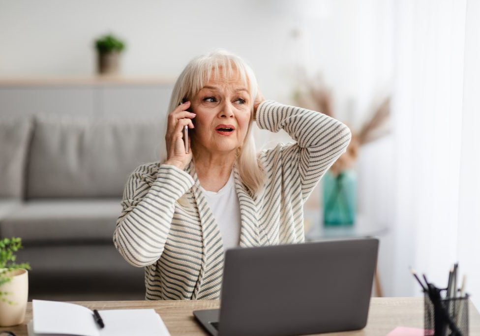 Oh No. Portrait of shocked frustrated middle-aged woman having problems with computer or work, talking on cellphone with customer support while working in home office grabbing head
